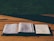 An open book and a notebook with handwritten notes sit on a wooden table under natural light. A pencil is placed in the book, suggesting it's being used for study or preparation.
