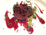A gourmet dish featuring succulent slices of beef topped with fresh herbs, accompanied by asparagus spears and a dark red sauce artfully spread on the plate.