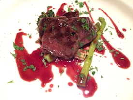 A gourmet dish featuring succulent slices of beef topped with fresh herbs, accompanied by asparagus spears and a dark red sauce artfully spread on the plate.