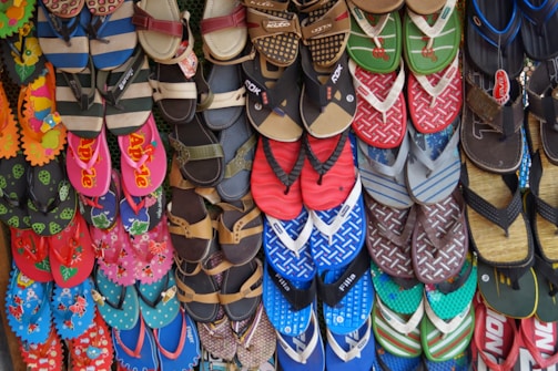 A colorful array of various flip-flops and sandals hanging on display. They feature multiple designs and patterns, including floral prints, stripes, and logos in a vibrant mix of colors like red, blue, green, and pink.