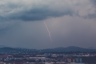 A dramatic scene with a single lightning bolt striking against a backdrop of dark, heavy clouds. The cityscape below features industrial buildings and structures, with distant hills visible in the horizon.