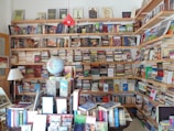 A cozy bookstore or personal library with wooden shelves filled with a vast array of books, from fiction to non-fiction. A globe sits prominently on a table, surrounded by more books and a lamp. The shelves are packed to the brim, indicating a love for literature and exploration.