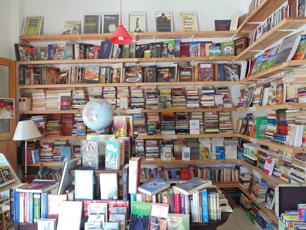 A cozy bookstore or personal library with wooden shelves filled with a vast array of books, from fiction to non-fiction. A globe sits prominently on a table, surrounded by more books and a lamp. The shelves are packed to the brim, indicating a love for literature and exploration.