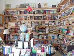 A cozy bookstore or personal library with wooden shelves filled with a vast array of books, from fiction to non-fiction. A globe sits prominently on a table, surrounded by more books and a lamp. The shelves are packed to the brim, indicating a love for literature and exploration.
