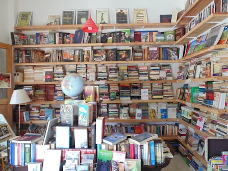 A cozy bookstore or personal library with wooden shelves filled with a vast array of books, from fiction to non-fiction. A globe sits prominently on a table, surrounded by more books and a lamp. The shelves are packed to the brim, indicating a love for literature and exploration.