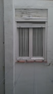 A closed white-framed window with vertical blinds is set into a light gray wall. The wall shows signs of aging and weathering, with some peeling paint above the window. Several thin lines or wires are attached to the window sill, stretching outward, possibly for drying clothes.