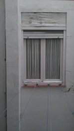 A closed white-framed window with vertical blinds is set into a light gray wall. The wall shows signs of aging and weathering, with some peeling paint above the window. Several thin lines or wires are attached to the window sill, stretching outward, possibly for drying clothes.