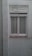 A closed white-framed window with vertical blinds is set into a light gray wall. The wall shows signs of aging and weathering, with some peeling paint above the window. Several thin lines or wires are attached to the window sill, stretching outward, possibly for drying clothes.