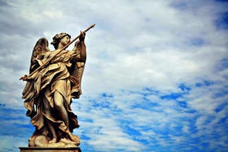 A detailed statue of an angel with wings, holding a spear-like object, set against a backdrop of a partly cloudy sky. The sculpted figure is intricately carved with flowing robes and appears to be standing majestically.