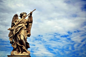 A detailed statue of an angel with wings, holding a spear-like object, set against a backdrop of a partly cloudy sky. The sculpted figure is intricately carved with flowing robes and appears to be standing majestically.