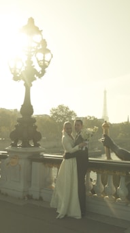 A couple in formal attire is posing on a decorative bridge framed by a lamppost with intricate details. The Eiffel Tower is visible in the background amidst a hazy, sunlit atmosphere. The woman holds a bouquet of flowers, and the couple appears to be dressed for a wedding.