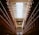 A corridor with multiple levels of cells stretching along both sides, likely within a prison. The perspective is from the middle of the corridor looking towards a bright skylight at the end of the hall. The cells have metal railings painted in red, and the corridor is mostly empty with minimal lighting from overhead lamps.