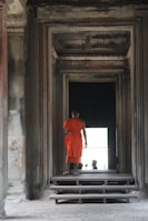 A person wearing an orange robe stands at the entrance of a dimly lit architectural structure, featuring intricate stone walls and steps leading inward.