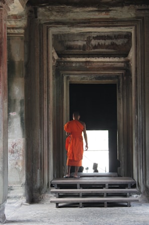 A person wearing an orange robe stands at the entrance of a dimly lit architectural structure, featuring intricate stone walls and steps leading inward.
