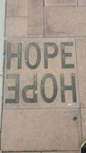 A sidewalk tile is inscribed with the word 'HOPE' in large, bold, black letters. The tiles have a textured appearance, and the surrounding tiles display various shades of brown and tan.