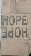 A sidewalk tile is inscribed with the word 'HOPE' in large, bold, black letters. The tiles have a textured appearance, and the surrounding tiles display various shades of brown and tan.