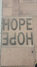 A sidewalk tile is inscribed with the word 'HOPE' in large, bold, black letters. The tiles have a textured appearance, and the surrounding tiles display various shades of brown and tan.