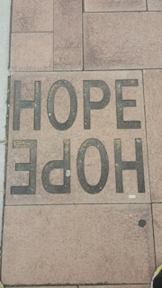 A sidewalk tile is inscribed with the word 'HOPE' in large, bold, black letters. The tiles have a textured appearance, and the surrounding tiles display various shades of brown and tan.