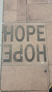 A sidewalk tile is inscribed with the word 'HOPE' in large, bold, black letters. The tiles have a textured appearance, and the surrounding tiles display various shades of brown and tan.
