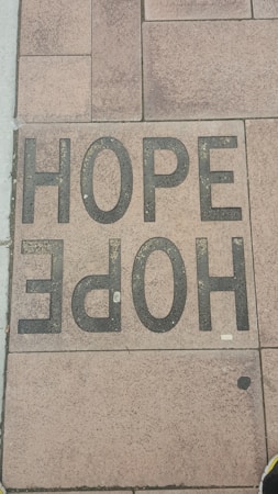 A sidewalk tile is inscribed with the word 'HOPE' in large, bold, black letters. The tiles have a textured appearance, and the surrounding tiles display various shades of brown and tan.