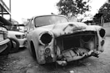 An old, rusted car without headlights or a grille is parked next to other vehicles in a lot. The car's exterior is heavily damaged with visible holes and peeling paint, reflecting years of neglect. The surrounding area is a mix of other cars, trees, and structures, creating an atmosphere typical of a junkyard or storage space.
