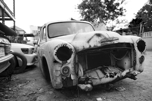 An old, rusted car without headlights or a grille is parked next to other vehicles in a lot. The car's exterior is heavily damaged with visible holes and peeling paint, reflecting years of neglect. The surrounding area is a mix of other cars, trees, and structures, creating an atmosphere typical of a junkyard or storage space.