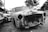 An old, rusted car without headlights or a grille is parked next to other vehicles in a lot. The car's exterior is heavily damaged with visible holes and peeling paint, reflecting years of neglect. The surrounding area is a mix of other cars, trees, and structures, creating an atmosphere typical of a junkyard or storage space.