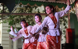 Three women dressed in traditional colorful costumes are performing a dance. They are wearing white blouses with red and gold patterned skirts and have their hair styled elegantly. The background features greenery and decorative elements, enhancing the cultural setting.