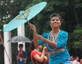 A person is performing a dance while elegantly holding a light blue parasol. The dancer is wearing traditional attire, which includes a blue garment and decorative jewelry. The background features people with umbrellas and lush greenery.