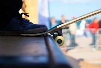 A close-up view of a skateboard positioned at the edge of a ramp with a person's foot resting on it. The focal point includes a black sneaker and the skateboard's wheels. The background is blurred, showing out-of-focus figures and a sunny, outdoor setting.