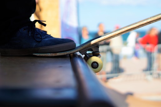 A close-up view of a skateboard positioned at the edge of a ramp with a person's foot resting on it. The focal point includes a black sneaker and the skateboard's wheels. The background is blurred, showing out-of-focus figures and a sunny, outdoor setting.