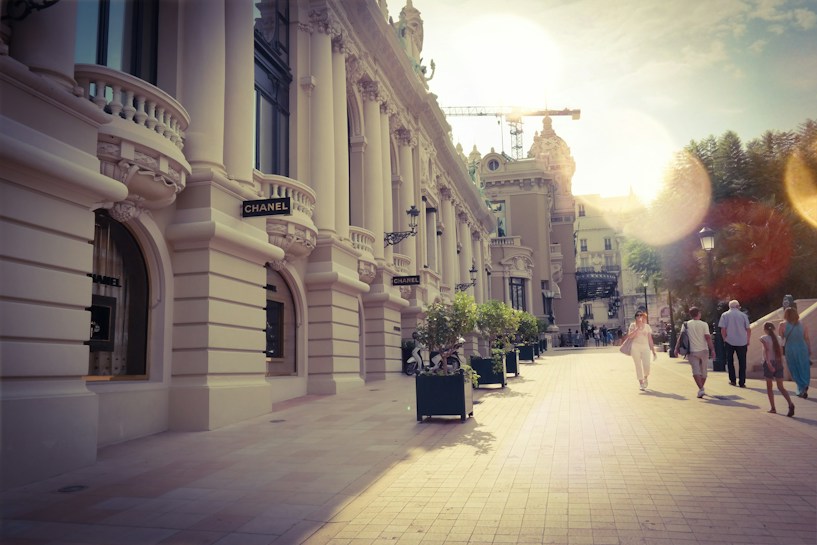 A stylish street scene with a luxurious building featuring ornate architecture and a Chanel storefront. Sunlight filters in, creating a warm and inviting atmosphere. People casually walk along the paved pathway, with trees and street lamps adding to the city's elegant ambiance.