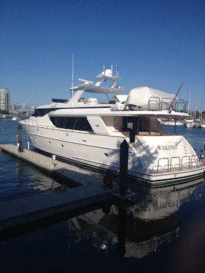 A luxurious white yacht docked at a marina under a clear blue sky. The yacht, named Viking, features multiple decks and modern amenities. Surrounding it are calm waters reflecting the vessel and the skyline of a nearby city with high-rise buildings.