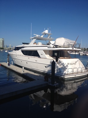 A luxurious white yacht docked at a marina under a clear blue sky. The yacht, named Viking, features multiple decks and modern amenities. Surrounding it are calm waters reflecting the vessel and the skyline of a nearby city with high-rise buildings.
