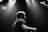 A black and white photograph featuring a musician on stage, illuminated by overhead spotlights. The individual is captured from the side, with a focus on their profile and upper body. The atmosphere is moody and dramatic, with soft light creating contrast against the dark background.