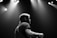 A black and white photograph featuring a musician on stage, illuminated by overhead spotlights. The individual is captured from the side, with a focus on their profile and upper body. The atmosphere is moody and dramatic, with soft light creating contrast against the dark background.