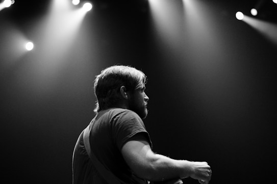 A black and white photograph featuring a musician on stage, illuminated by overhead spotlights. The individual is captured from the side, with a focus on their profile and upper body. The atmosphere is moody and dramatic, with soft light creating contrast against the dark background.
