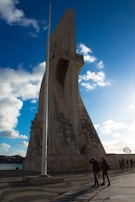 A large stone monument with intricate carvings stands against a bright blue sky partially covered with scattered clouds. People walk along a paved path beside the monument, and there is a large white flagpole adjacent to the structure. The scene is bathed in late afternoon light, casting shadows on the monument and ground.