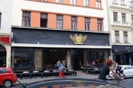 A street view of a café with a dark facade and gold emblem above the entrance. People are seated outside at small round tables, and a person on a bicycle is passing by. The building has large windows with red frames and beige upper floors.