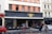 A street view of a café with a dark facade and gold emblem above the entrance. People are seated outside at small round tables, and a person on a bicycle is passing by. The building has large windows with red frames and beige upper floors.