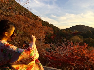 A person wearing a colorful kimono is taking a photograph with a camera in their hands. The scene is set during autumn, featuring vibrant red and orange foliage. A distant mountain and pagoda are visible under a clear sky, providing a serene and picturesque backdrop.