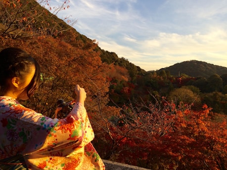 A person wearing a colorful kimono is taking a photograph with a camera in their hands. The scene is set during autumn, featuring vibrant red and orange foliage. A distant mountain and pagoda are visible under a clear sky, providing a serene and picturesque backdrop.