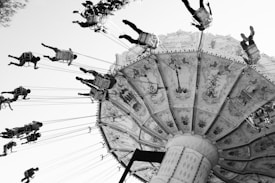 A large, intricately decorated carousel swing ride is in motion. The swings are suspended on long chains, and numerous people are seated, flying outward due to centrifugal force. The image is in black and white, highlighting the contrast between the detailed patterns on the ride and the clear sky.