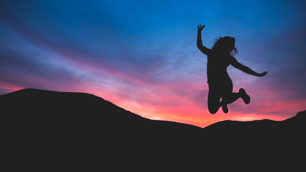 A woman in silhouette jumps for joy against dark mountains and a pink and purple sunset.