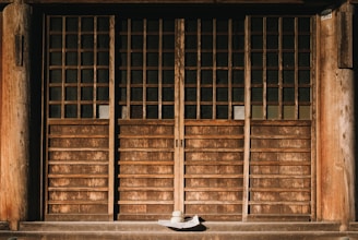 A traditional wooden door with wooden lattice design and paneling, accompanied by a wide-brimmed hat placed on the ground in front. The door has a rustic and weathered appearance, reflecting age and history. The shadows cast by the sunlight add depth and highlight the texture of the wood.