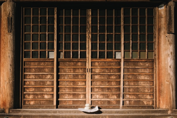 A traditional wooden door with wooden lattice design and paneling, accompanied by a wide-brimmed hat placed on the ground in front. The door has a rustic and weathered appearance, reflecting age and history. The shadows cast by the sunlight add depth and highlight the texture of the wood.