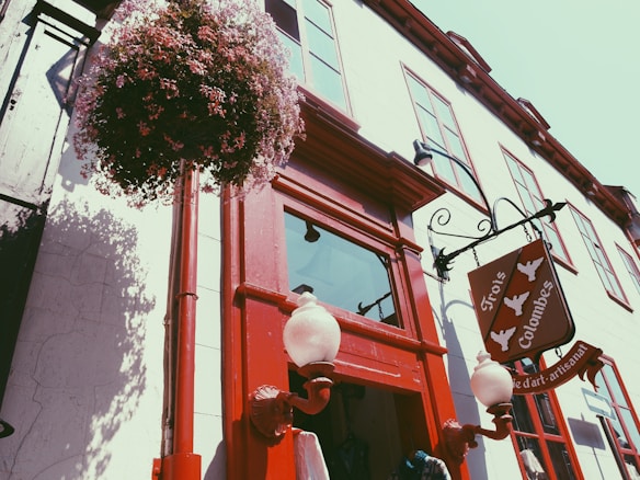 A picturesque street scene featuring a traditional building with red-framed windows and a red door. A decorative wrought iron sign hangs above the entrance, displaying the name 'Trois Colombes' alongside three dove silhouettes. A large hanging flower basket adds a touch of nature, while ornate street lamps provide additional detail.