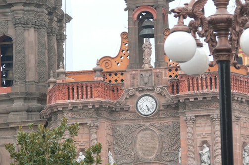 A detailed architectural structure features ornate carvings and a clock with a classical design. Statues are integrated into the building's facade, adding to its historical and artistic significance. The material appears to be stone with intricate detailing and rustic coloring. In the foreground, a vintage-style street lamp with multiple round bulbs adds to the scene. Lush green foliage can be seen at the bottom, providing a natural contrast to the stone architecture.