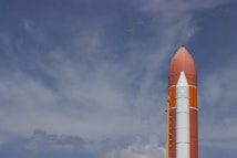 A large rocket standing upright against a partly cloudy sky, showcasing its pointed nose and external fuel tank.