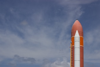 A large rocket standing upright against a partly cloudy sky, showcasing its pointed nose and external fuel tank.